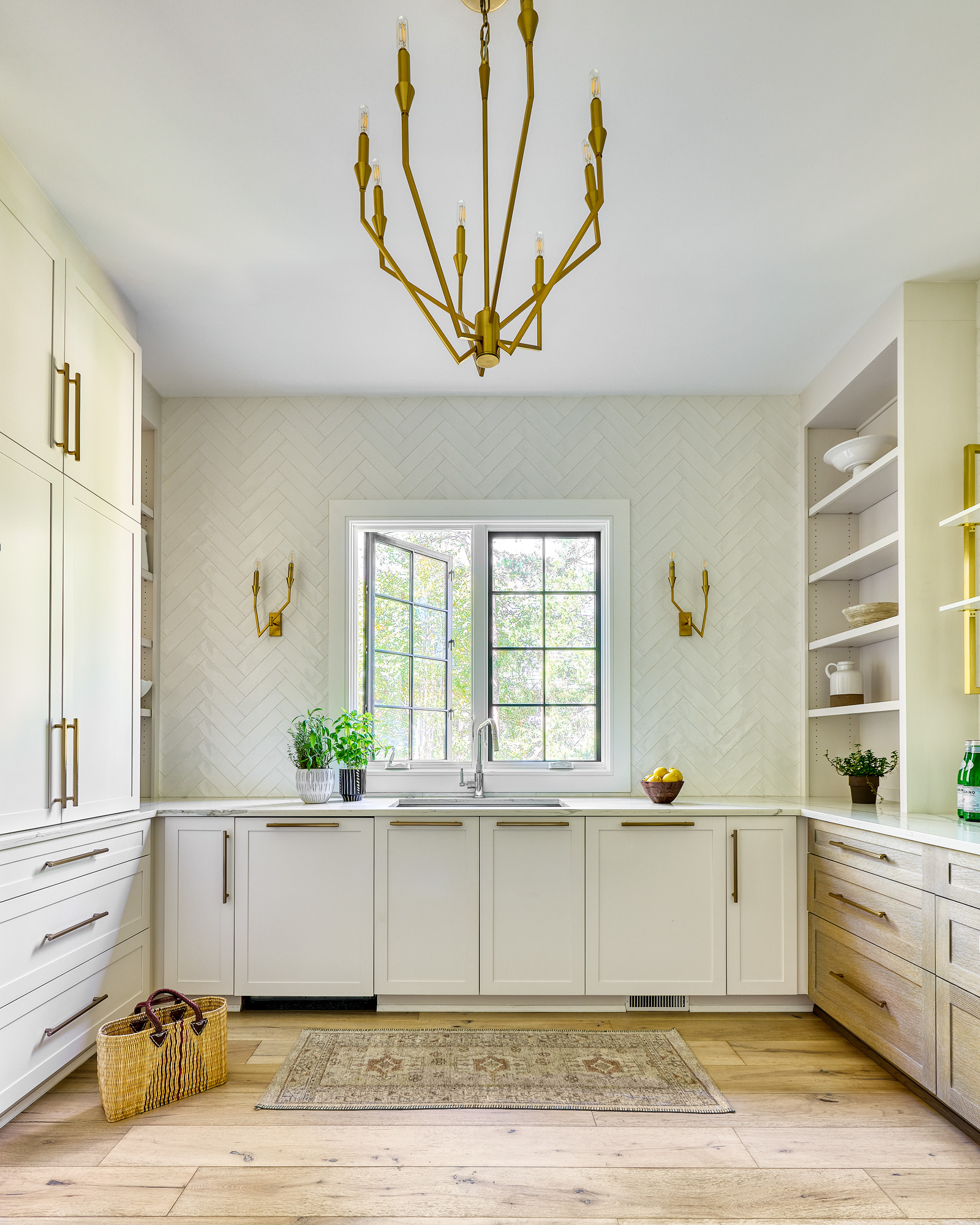 Butler prep-kitchen inside a Highgate estate — herringbone tile backsplash, brass chandelier, honed marble counters, white and rift-oak cabinetry