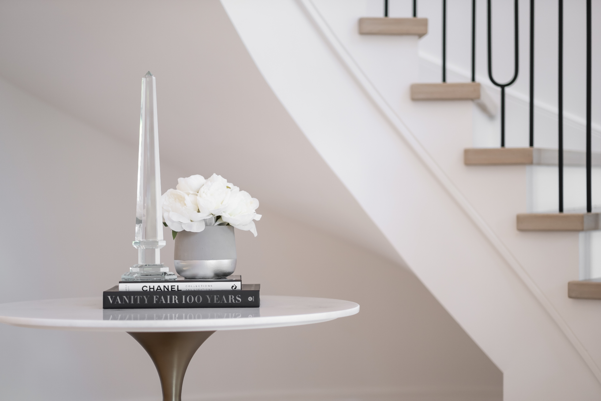Foyer vignette inside a Highgate estate — crystal obelisk and white peonies on a marble tulip table beneath a curved staircase with iron balusters